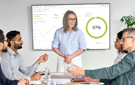 Smiling woman talking to people around a table. Behind her a screen with Unit4 FPA