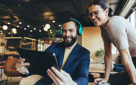 A man in a blue jacket and turquoise headphones smiles while using a tablet, with a woman beside him