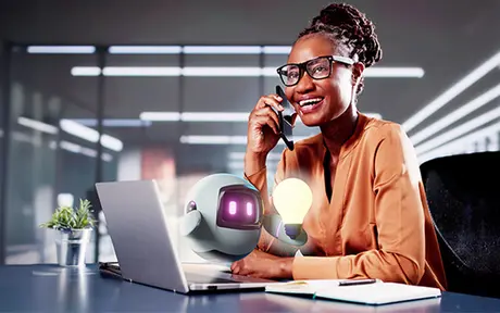 A woman smiling on a phone while sitting at a desk with a laptop and a cartoon robot holding a light bulb. 