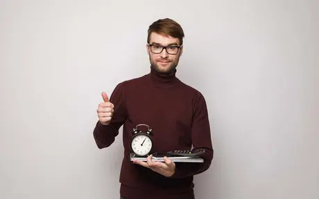 A man in a burgundy turtleneck holds an alarm clock, a calculator, and notebooks, giving a thumbs-up.