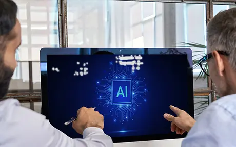Two individuals collaborating in an office, focusing on a computer screen displaying a digital circuit board with 'AI' prominently featured, symbolizing artificial intelligence and teamwork.