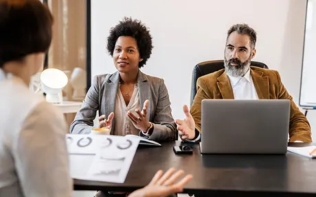 Two business people in suits listening to a presenter in a meeting room.
